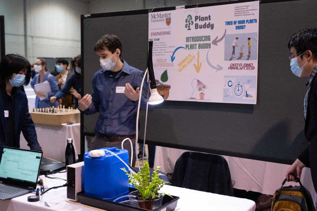 Plant Buddy Capstone display, a plant sits on a table with a light shining on it and a water jug beside it. It's connected to a computer that tracks when and how to take care of it.