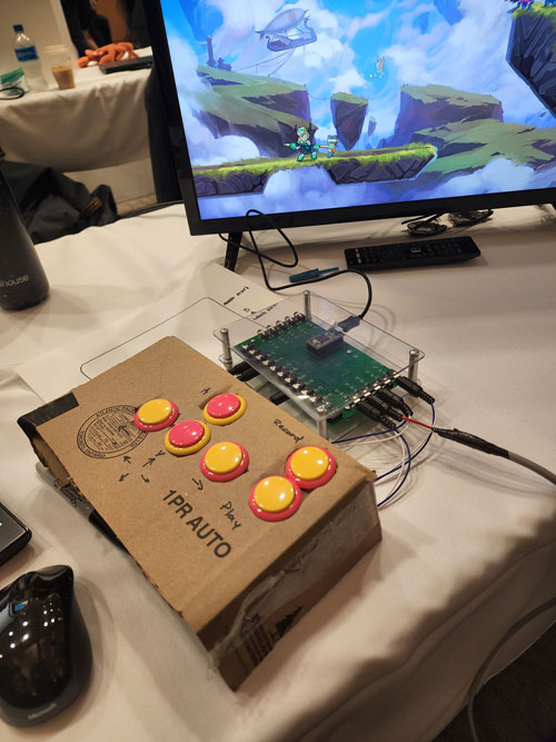 Accessible Game Controller capstone display: a cardboard prototype with pink and yellow buttons is connected to a circuit board and computer that shows a video game playing in the background.