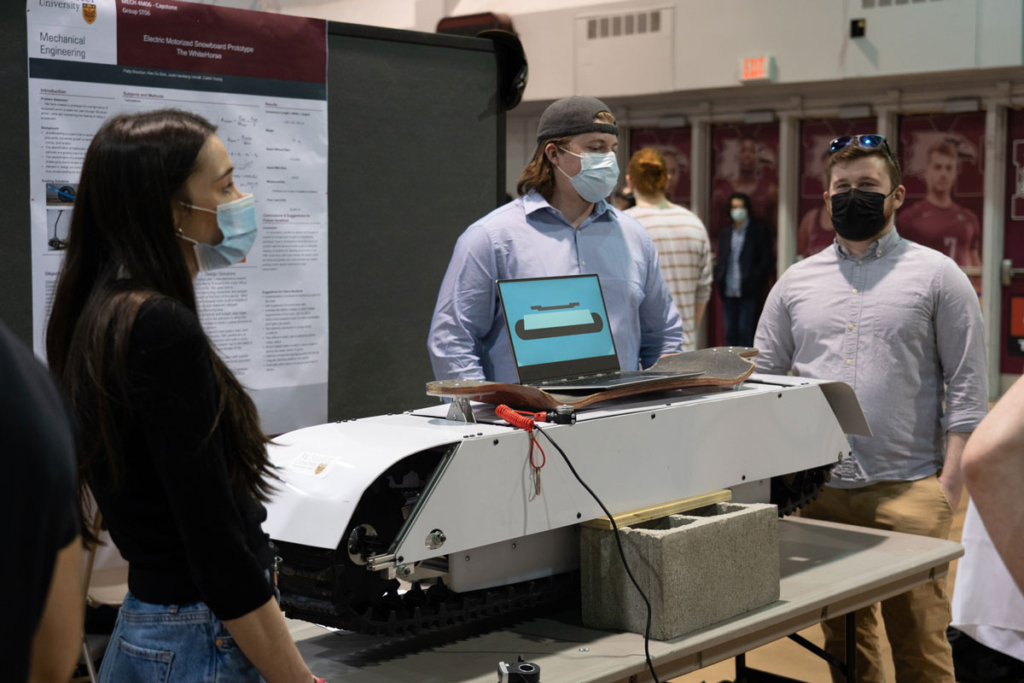 Members of the Electric motorized snowboard capstone pose with their prototype: a long white board with treads and a skateboard on top of it.