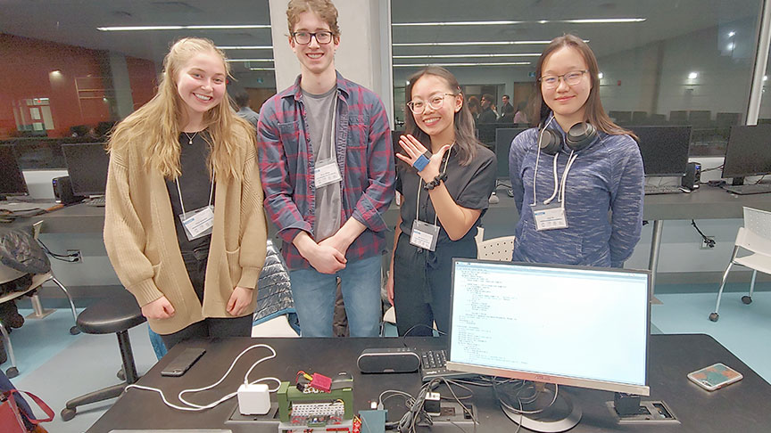 four students stand behind a computer with a device on one of their hands