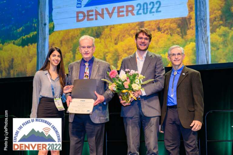 John Bandler stands on stage with three others at the closing session of the International Microwave Symposium in 2022.