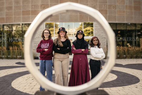 Four women stand behind the iron ring in front of the John Hodgins Engineering Building.