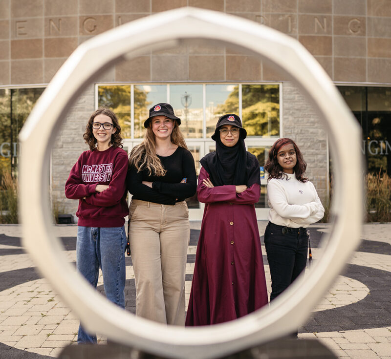 Four women stand behind the iron ring in front of the John Hodgins Engineering Building.
