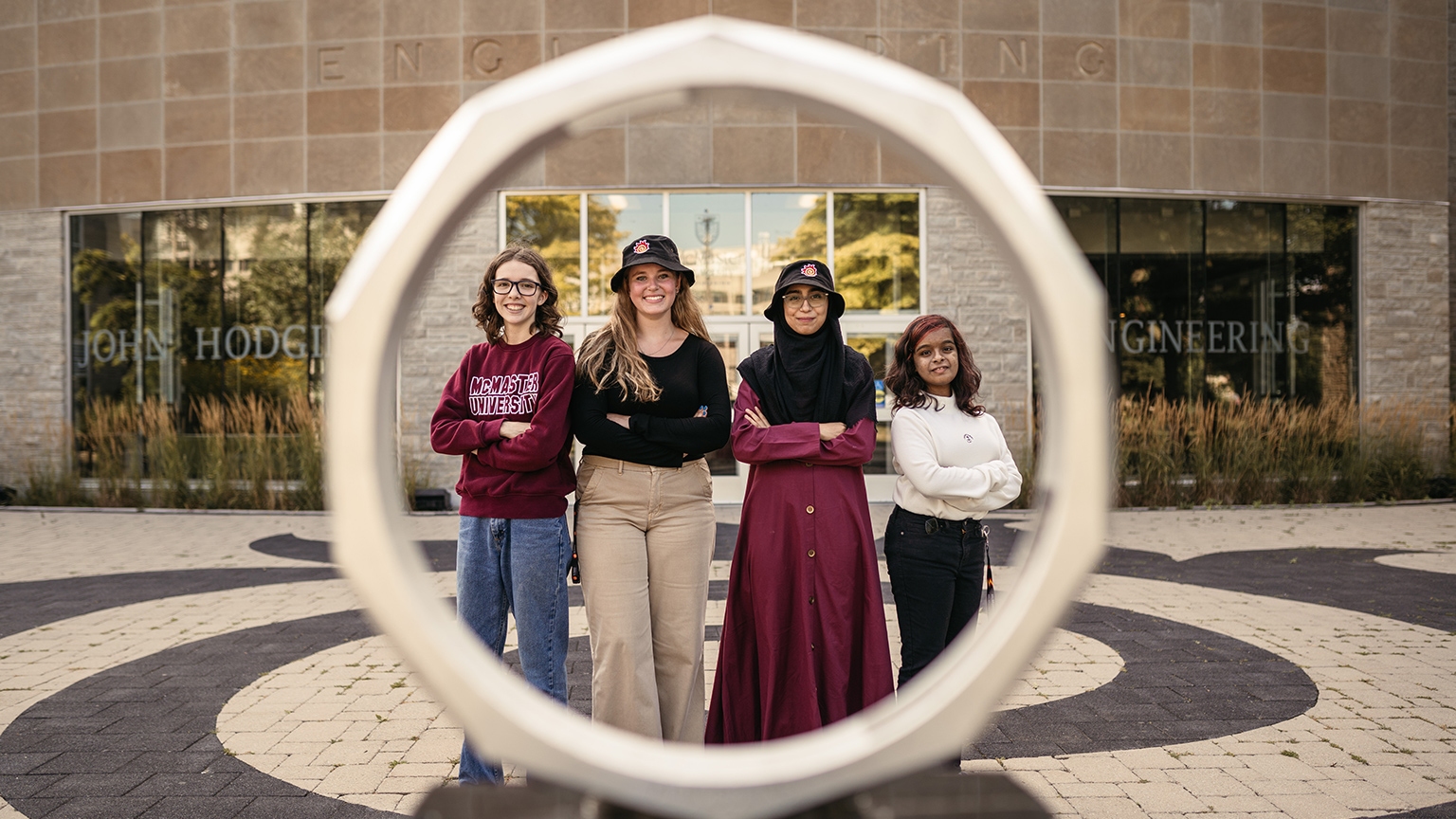 Four women stand behind the iron ring in front of the John Hodgins Engineering Building.