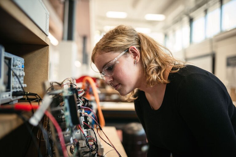 Chelsey Ellise wears safety googles as she leans forward to look at some wires on the left.