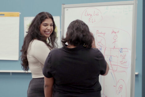 Two students stand at a whiteboard that has words and drawings on it. One student has their head turned and is smiling at the other student whose back is turned to the camera.