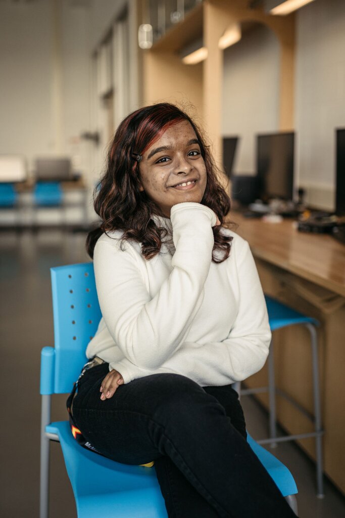 Neaha Bijo sits on a chair, smiling with her hand on her chin.