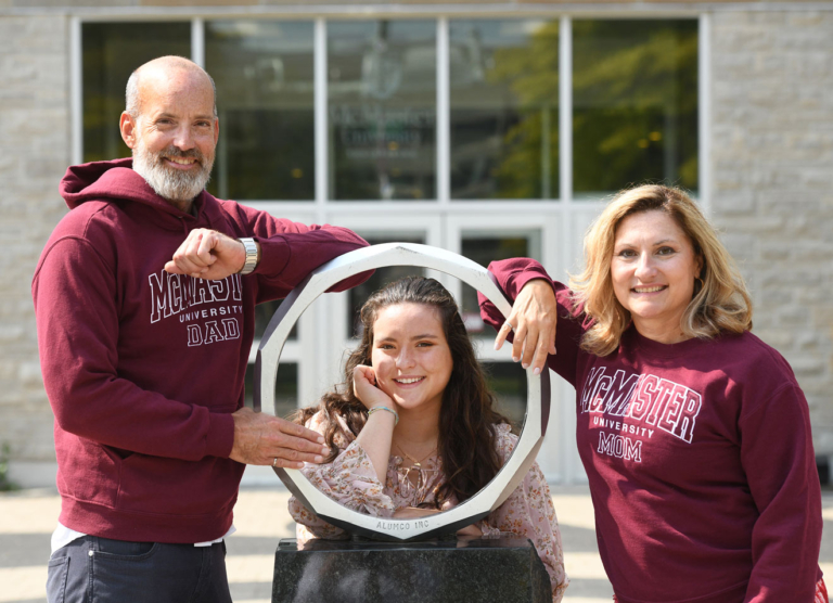 Two grads wearing McMaster mom and dad sweaters are on each side of the iron ring statue while their child poses in the ring
