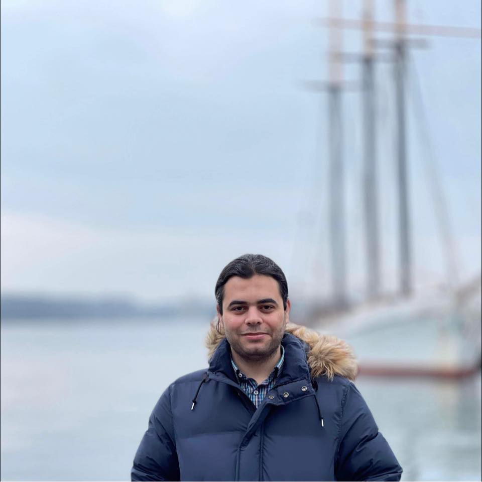 Ahmed Moussa poses in front of a body of water with a three mast boat in the background
