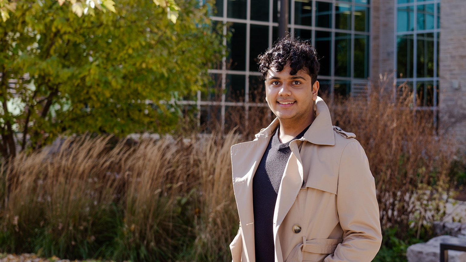 Yuvi Jain poses in a tan trench coat in front of JHE