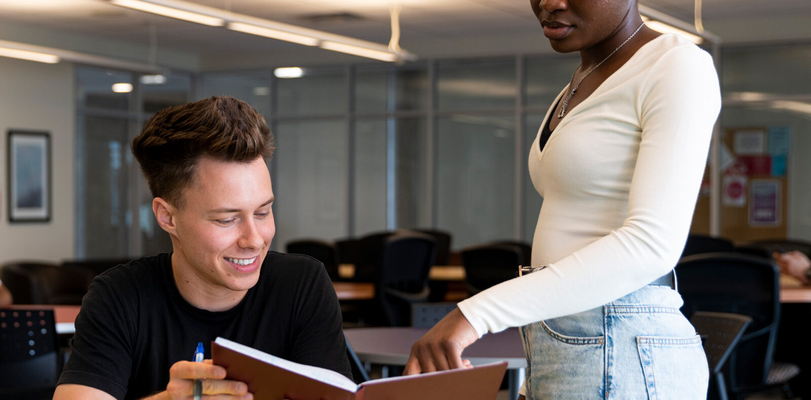 A student sits at a table with an open book. Another student stands beside them and points at a page.