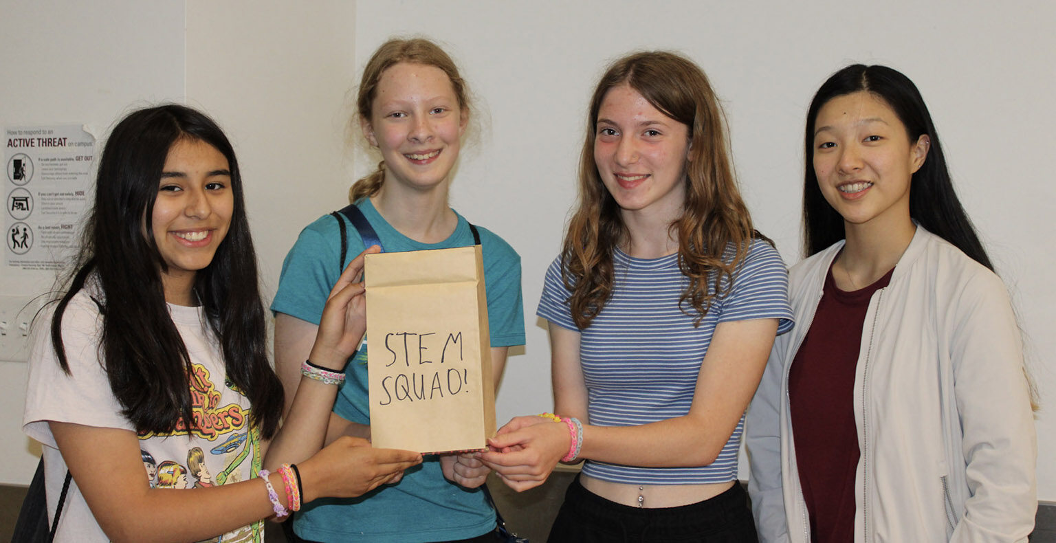 Four girls holding up a paper bag with the words STEM Squad written on it