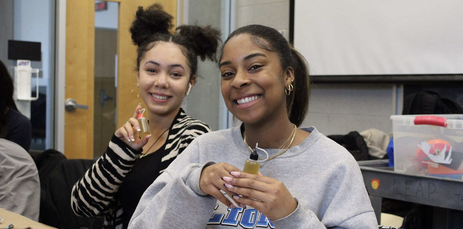 Two young women smiling, holding hair oil containers that they created in a STEM program