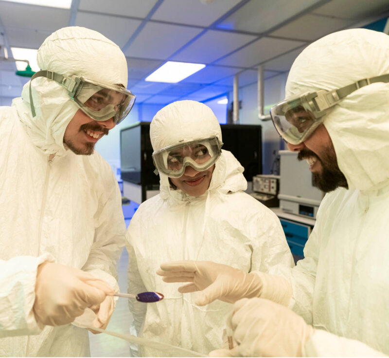 Three people in protective gear talk over a petri dish.