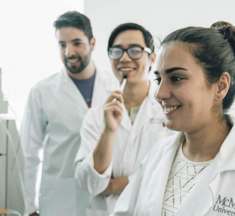 A woman in a McMaster lab coat smiles with two other people.