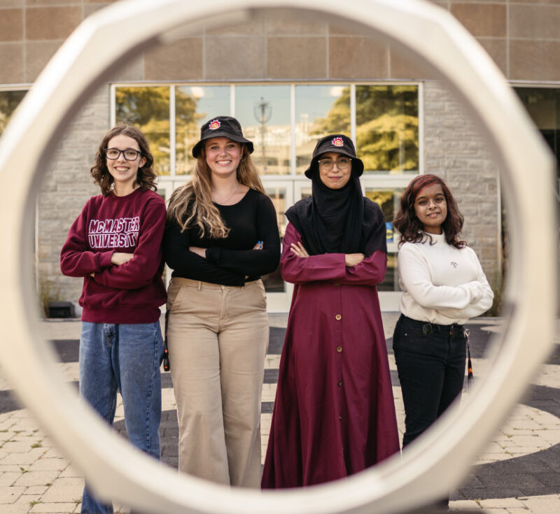 Four women students standing in front of the iron ring on campus