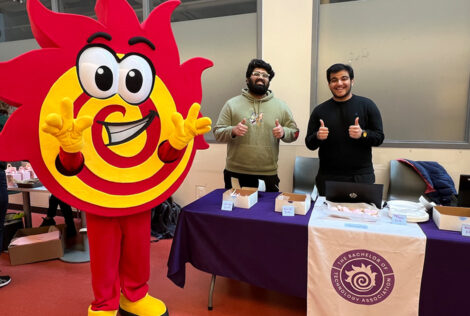 Flare the Fireball mascot stands beside Gopal and Rumman, who are running a bake sale.