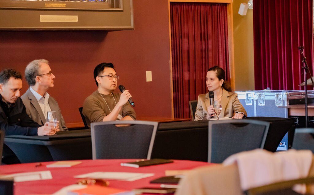 four people sitting at a long black table as part of a panel. One of them is holding up a microphone and speaking into it.