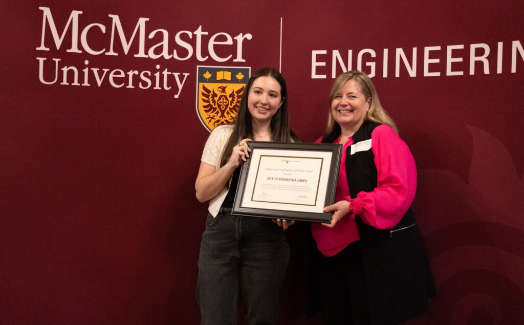 two women standing in front of a McMaster University sign holding an award.