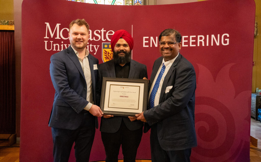 three men standing in front of a McMaster University sign holding an award.