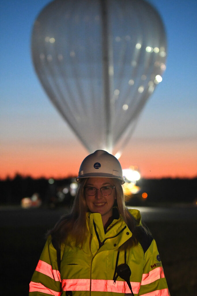 Magalie wears a hard hat and poses in front of a stratopheric balloon with a beautiful sunrise in the background