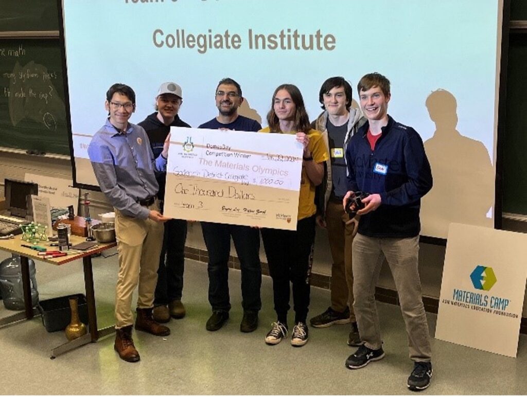 a group of students standing at the front of the room, in front of a projector that has a presentation on it, being awarded a large check by a professor.