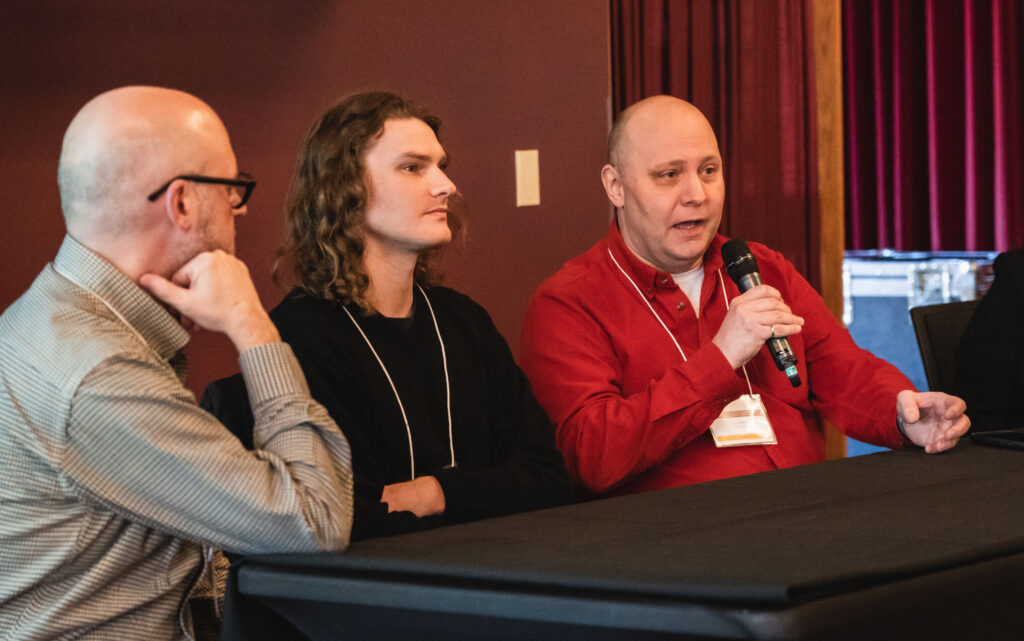 three people sitting at a long black table as part of a panel. One of them is holding up a microphone and speaking into it.