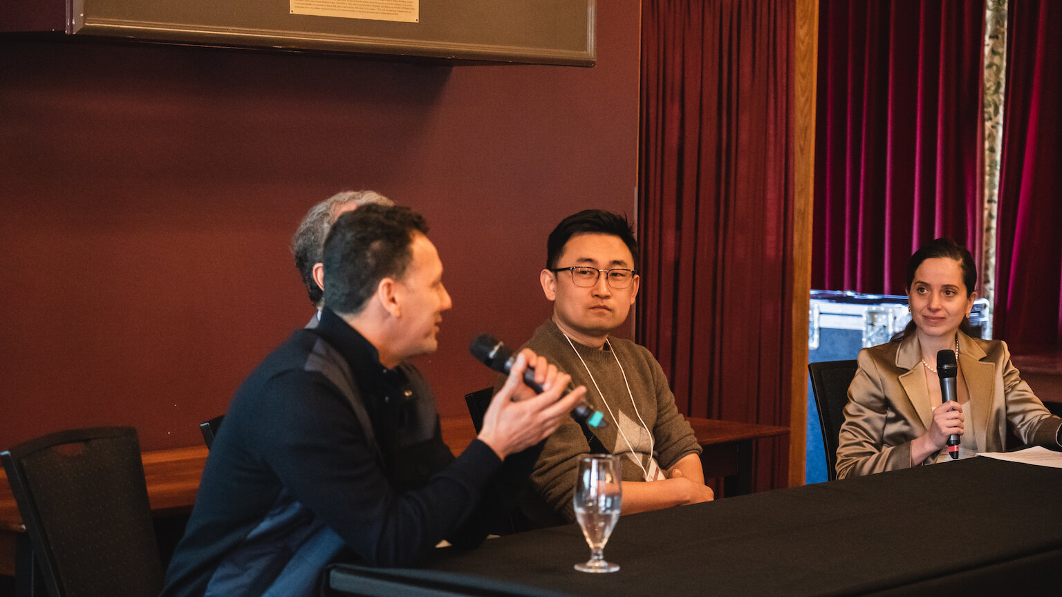 four people sitting at a long black table as part of a panel. One of them is holding up a microphone and speaking into it.