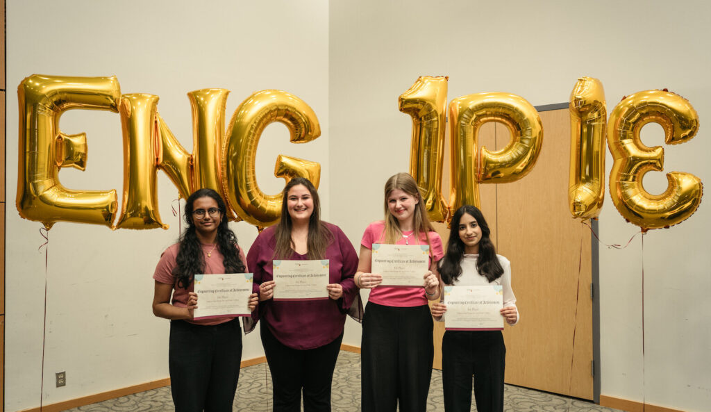 Four people standing in front of gold foil balloons holding up certificates.