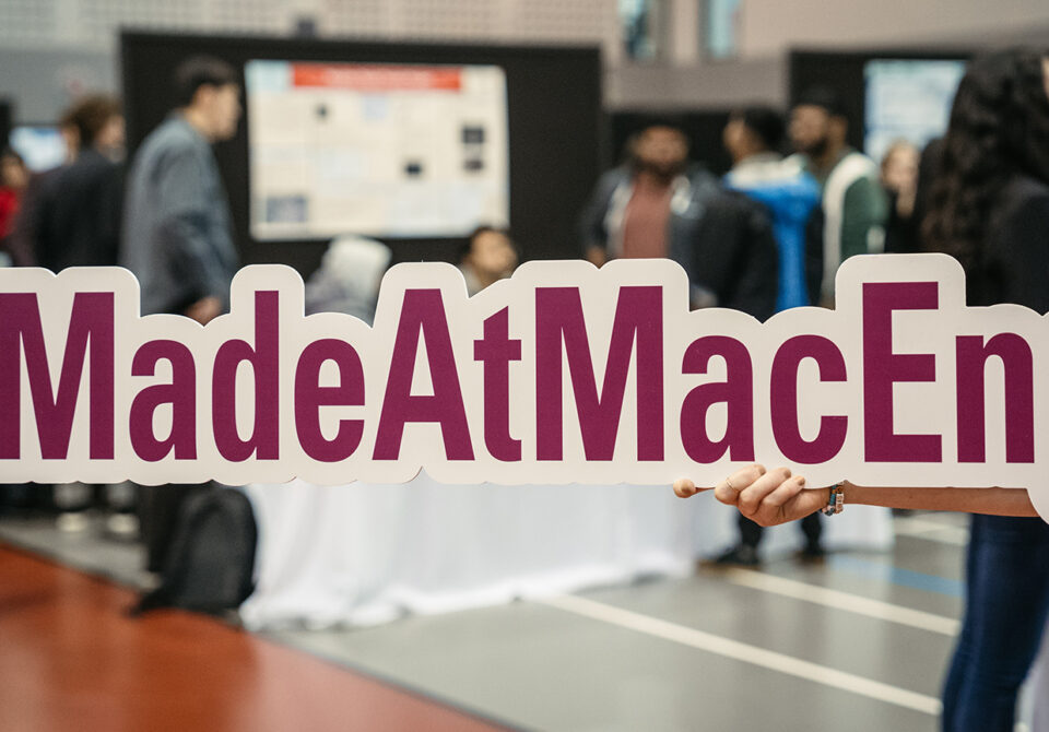 A #MadeAtMacEng sign is held up with the hustle and bustle of Capstone Expo going on behind it