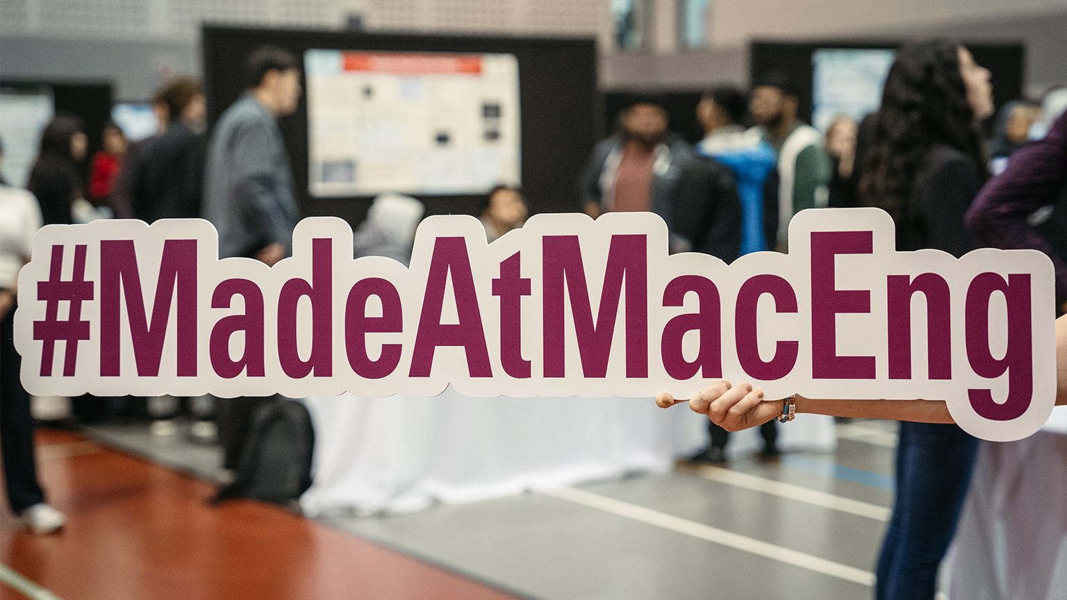 A #MadeAtMacEng sign is held up with the hustle and bustle of Capstone Expo going on behind it