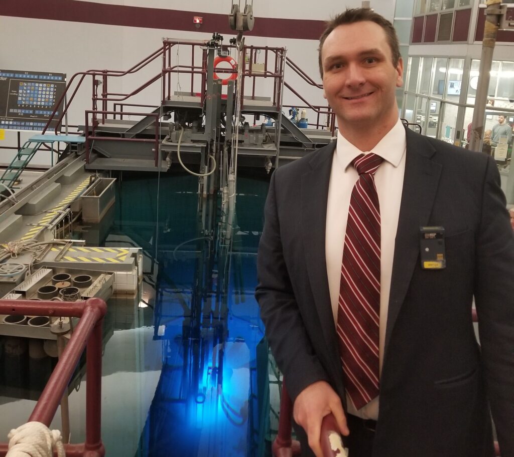 man standing in a nuclear reactor, with the view of a blue light core in a swimming pool showing behind him.