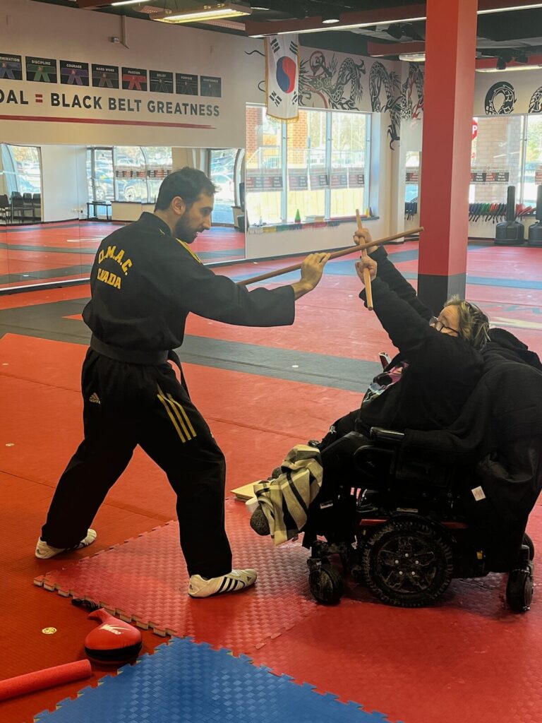 a woman in a wheelchair sparring with a martial arts teacher at a martial arts studio.