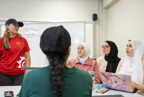 an instructor in a red t-shirt speaking to a group of four women sitting at a desk.
