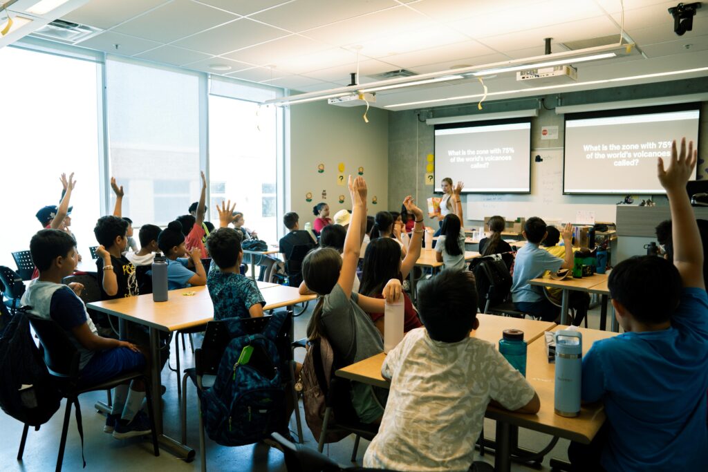 young camp students in classroom with their hands raised as they participate in a workshop