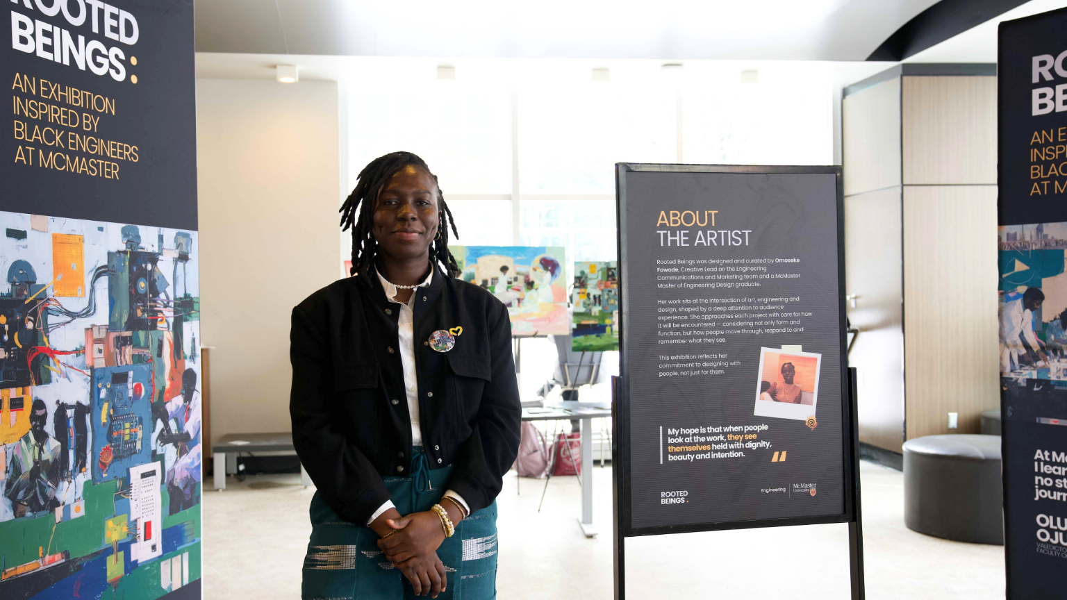 Artist Omoseke Fowode standing in front of the Rooted Beings exhibition in JHE Lobby on campus