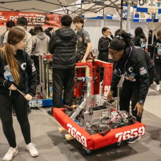 A team at a FIRST Robotics event moves their robot through a busy pit area as other teams work on their machines nearby.