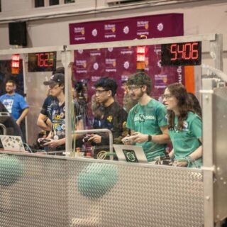 Drivers from multiple teams stand at the control station operating their robots during a FIRST Robotics match, with score displays and field barriers visible in front of them.
