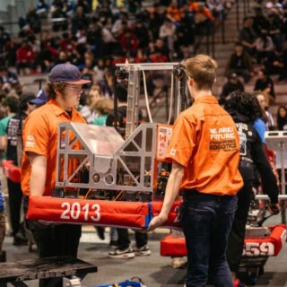 Two team members in orange shirts carry their robot labeled 2013 across the competition floor during a FIRST Robotics event.