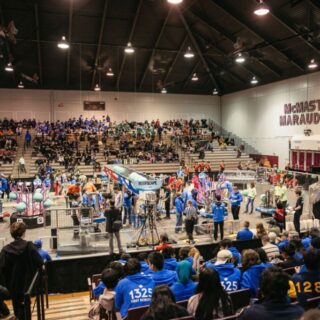 A large crowd watches as teams and referees gather around the competition field during a FIRST Robotics event inside a busy arena.
