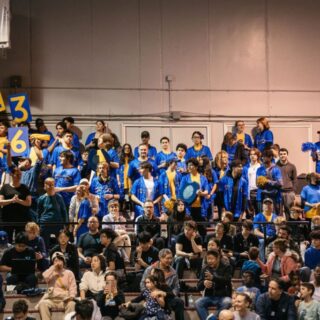 A large group of audience in blue and yellow team colors cheer from the stands during a FIRST Robotics event.