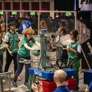 Team members in green gather around their robot, labeled 5406, as they prepare it on the field during a FIRST Robotics competition match.