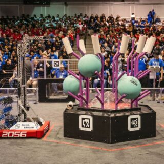 A FIRST Robotics competition field with a scoring structure holding large teal game pieces, as a robot from Team 2056 approaches while a packed audience watches from the stands.