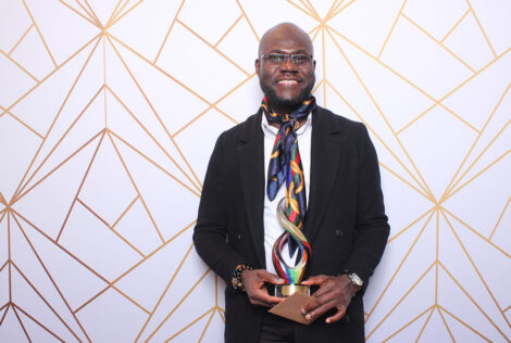 Albert Ashong holds a colourful award that matches his colourful scarf in front of a white background with a funky gold pattern