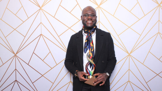 Albert Ashong holds a colourful award that matches his colourful scarf in front of a white background with a funky gold pattern