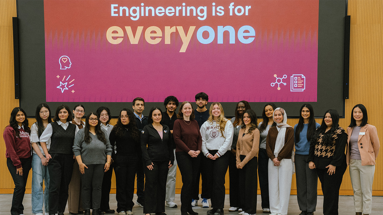 A group of women and men whose work at Mac Eng positively influences the lives of women pose together in front of a screen that reads engineering is for everyone