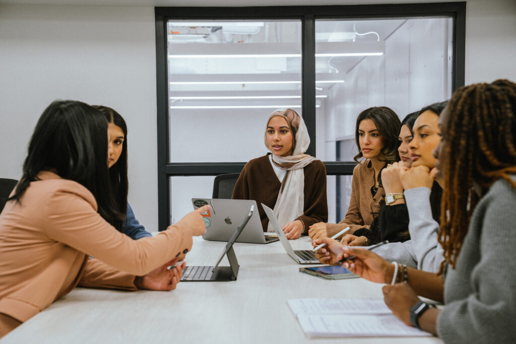 Zoe talks to the members of the capstone group at a table, a few of them have their hands under their chins focused on taking what Zoe is sharing in