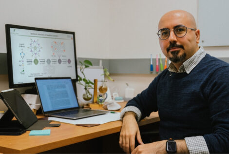 Reza sits at his desk looking over straight into the camera, a laptop sits open in front of a monitor that shows his work