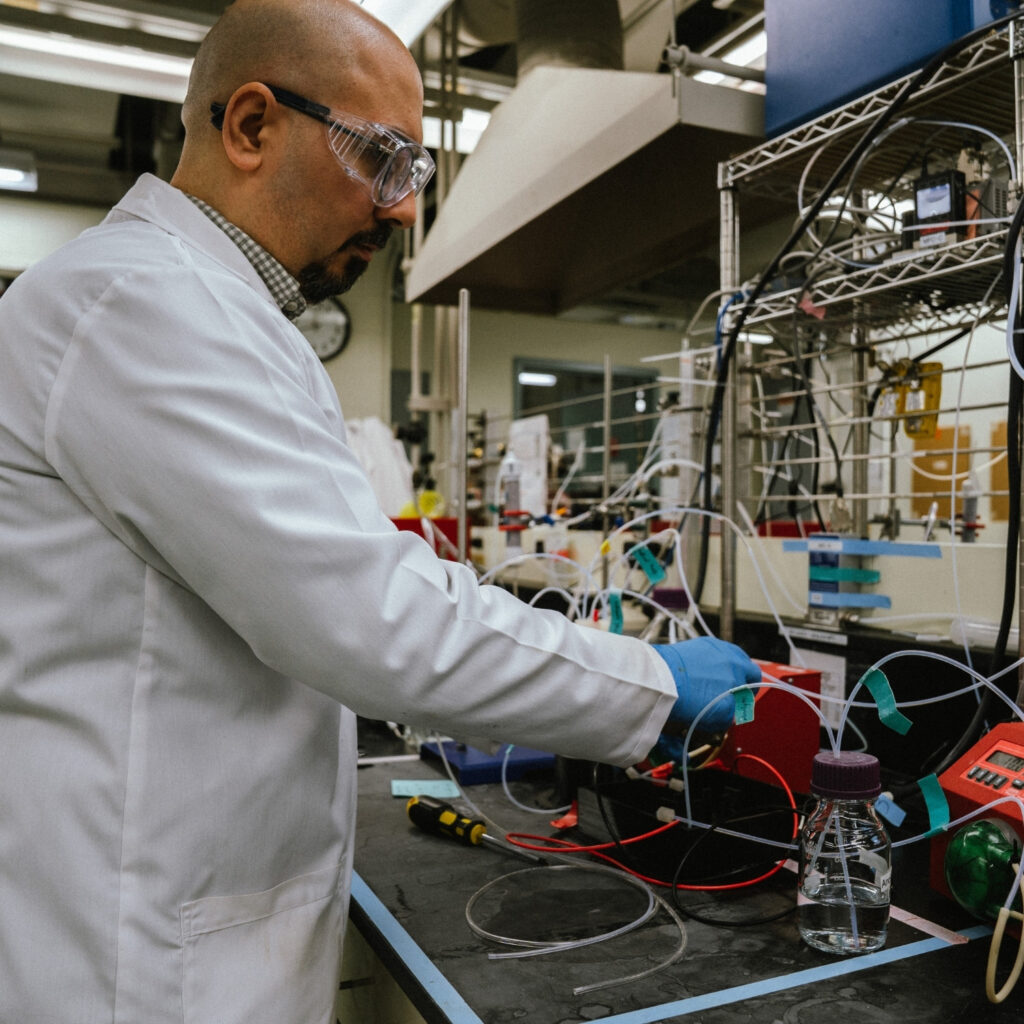 Reza in the lab wearing a coat working at the counter with tubes leading from a small bottle filled with liquid