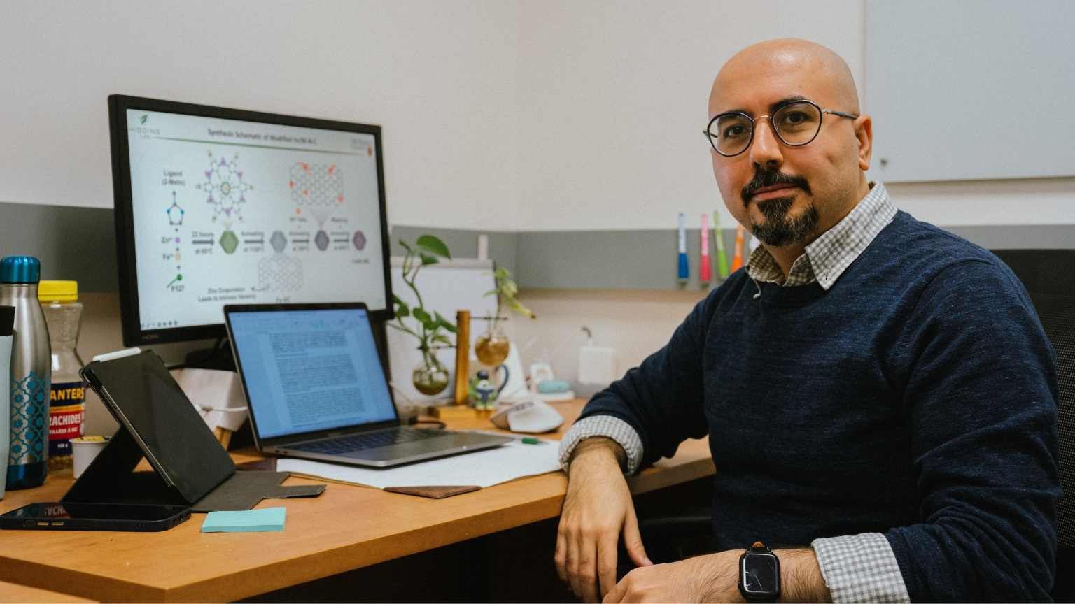 Reza sits at his desk looking over straight into the camera, a laptop sits open in front of a monitor that shows his work